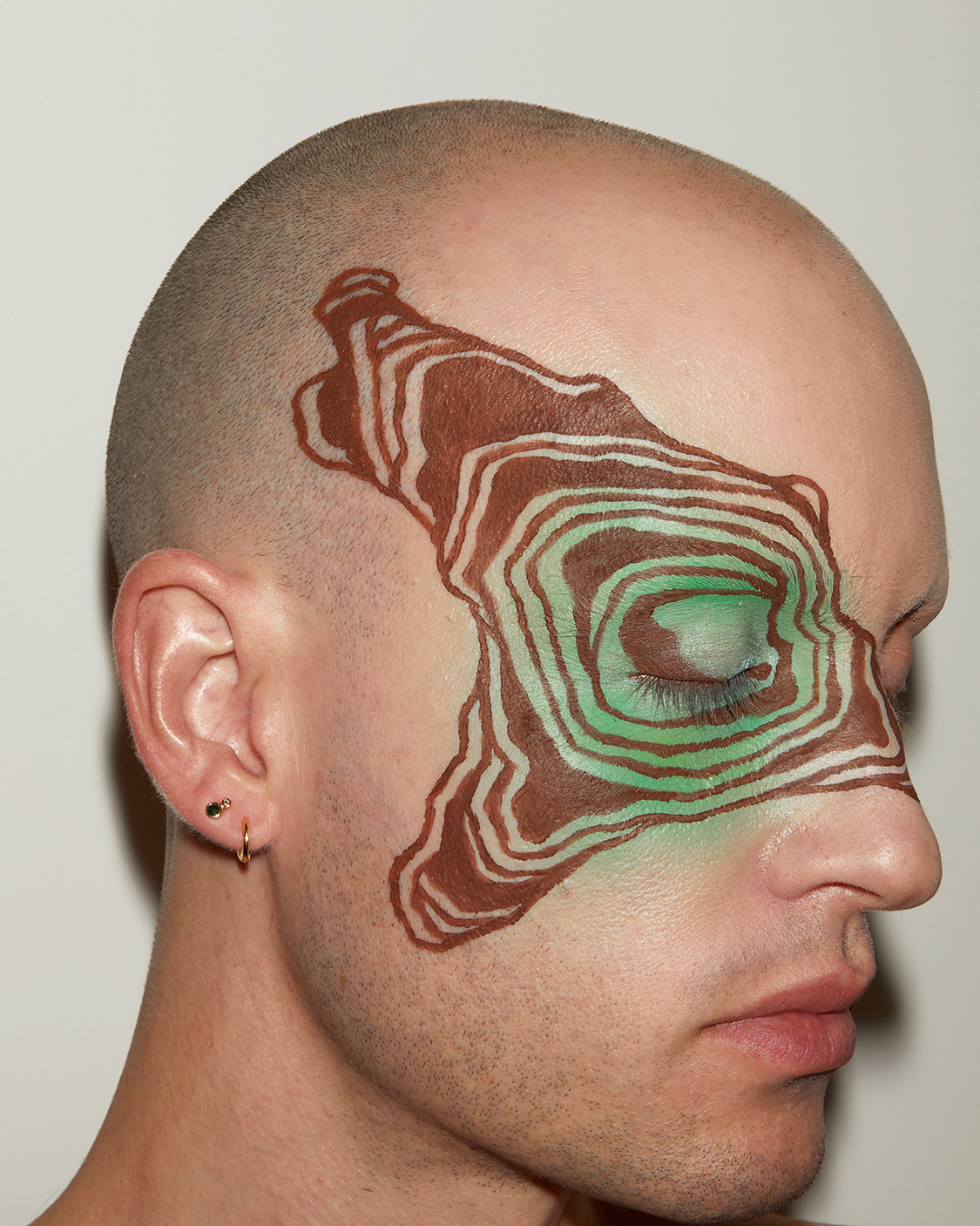 Close up portriat of Jesse in profile with graphic brown liner make up in concentric shapes extending out from one eye against a grey background